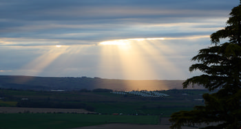 This landscape photograph captures the striking sight of God's rays, also known as crepuscular rays, streaming through a break in the clouds on a spring evening over the rural countryside near Bolsover in Derbyshire, United Kingdom. The sunlight pierces the cloudy sky, illuminating a solar farm with rows of solar panels set across the rolling green fields of this rural part of England. The dramatic interplay of light and shadow emphasizes both the beauty of the natural landscape and the presence of sustainable energy technology in the region. The image perfectly illustrates the unique atmosphere of a spring evening in the English countryside, with clouds diffusing the fading sunlight and casting long rays across the landscape.